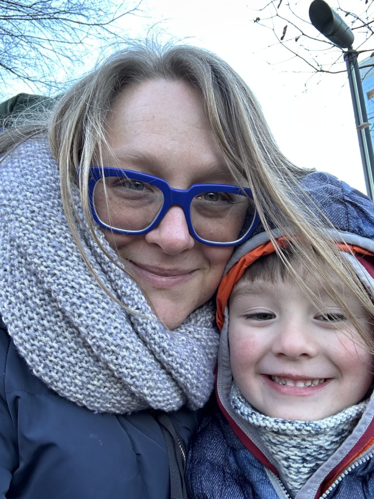 A picture of a white woman wearing blue glasses and a preschool aged boy. They are both bundled in winter clothes and are outside. 